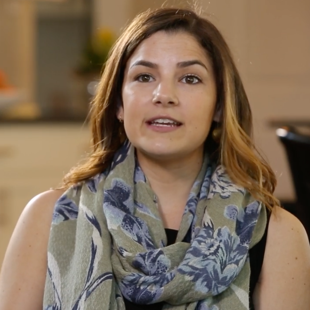 A woman with light brown hair, wearing a black top and a large blue and green floral scarf, sits indoors with a neutral expression. A kitchen background is softly blurred behind her.
